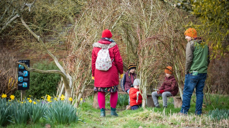 A family sitting on a bench under a tent-like structure weaved out of willow in the walled garden.